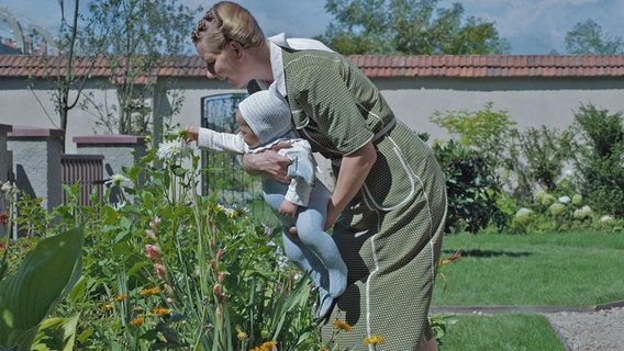 Sandra Hüller (2.v.r.) als Mutter der Familie Höss hält im Outfit der 40er-Jahre mit hochgesteckten Haaren ein Baby mit Haube in ein Blumenbeet - Szene aus dem Spielfilm "The Zone of Interest" von Jonathan Glazer © Leonine 