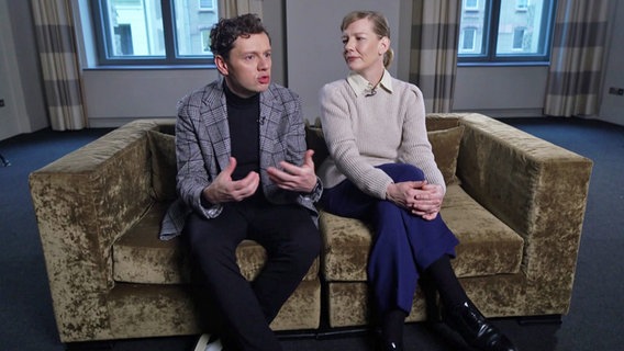 Christian Friedel und Sandra Hüller sitzen auf einem grau-braunen Sofa. © NDR 