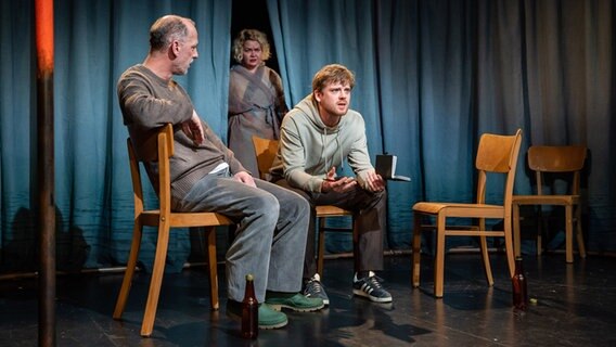 Zwei Darsteller sitzen auf Stühlen, während eine dritte Darstellerin hinten durch einen Vorhang auf die Bühne tritt. © Schleswig-Holsteinisches Landestheater 