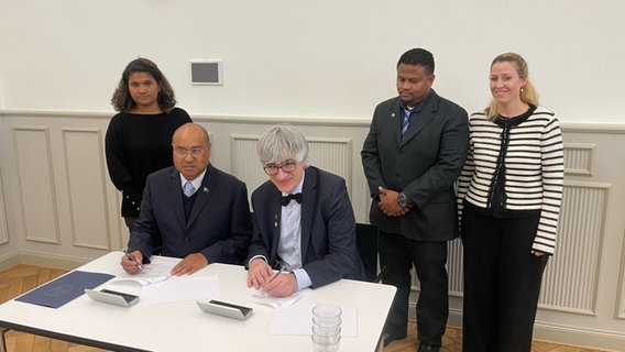 Ngiraibelas Tmetuchl (Kulturminister aus Palau) und Metin Tolan (Präsident der Uni GÖ) v.l. sitzen an einem Tisch und unterschreiben einen Vertrag. © NDR Foto: Doretta Farnbacher