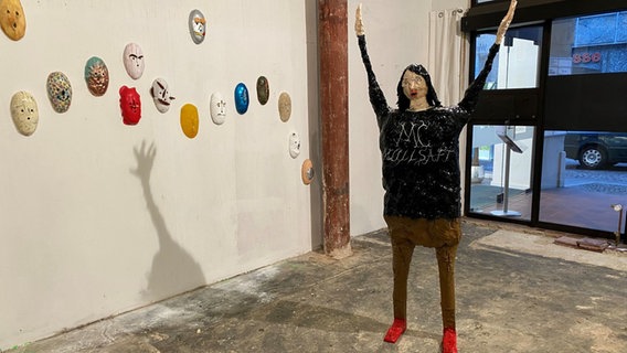 Eine Figur steht mit erhobenen Armen mitten in einem Raum, an der Wand hängen bunte, ovale Masken. © Linda Ebener / NDR Foto: Linda Ebener