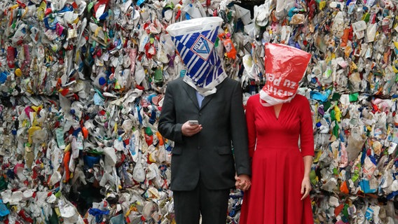 Zwei Menschen mit Plastiktüten über dem Kopf stehen vor einem Plastikmüllberg © Swaantje Güntzel/Scheibe & Güntzel Foto: Scheibe & Güntzel