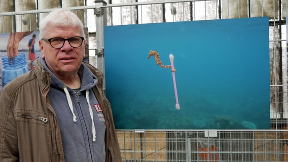 Claus Spitzer-Ewersmann vor einem Bild in der Ausstellung "Planet or Plastic?", das ein Seepferdchen zeigt, welches mit seinem Schwanz einen Q-Tip umschlugen hält. © NDR 