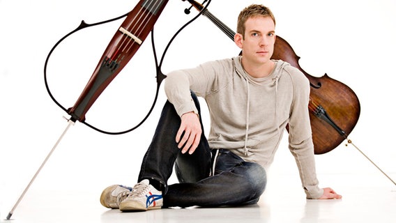 Der Cellist Johannes Moser sitzt vor zwei Instrumenten, einem akkustischem und einem elektrischem Cello.  