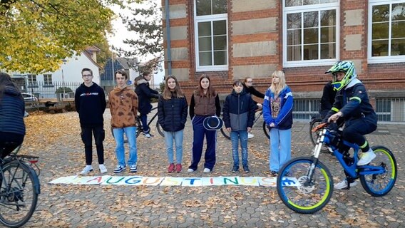 Sechs Schülerinnen und Schüler stehen auf dem Schulhof und werden von Kindern auf Fahrrädern umkreist. Vor ihnen liegt Papier mit der Aufschrift "Augustinus" © NDR 