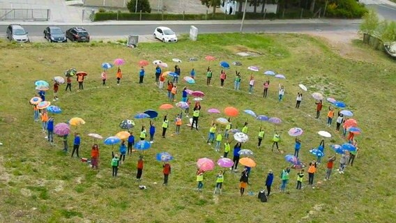 Etwa sechzig Menschen stehen in Form eines Peace-Zeichens auf einer Wiese und halten Regenschirme in die Luft © NDR 