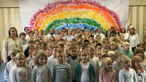 Etwa sechzig Kinder stehen mit ein paar Erwachsenen vor einem riesigen Regenbogen aus Handabdrücken © NDR 