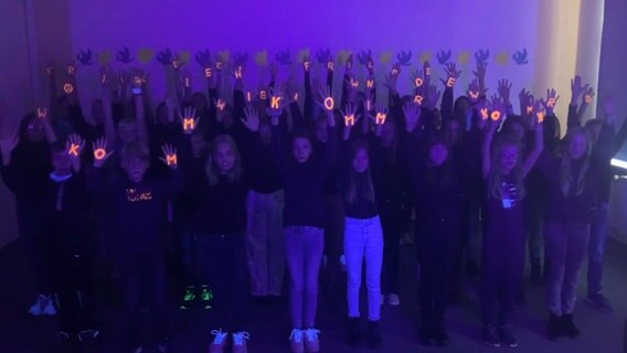 Etwa dreißig Kinder stehen bei Schwarzlicht zusammen und halten ihre Hände in die Luft, auf der einzelne Leuchtbuchstaben den Text "Komm, wir zieh'n in den Frieden" bilden © NDR 