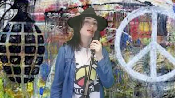 Eine junge Frau in T-Shirt und Jeanshemd mit schwarzem, breitkrempigen Hut hält ein Standmirkofon in der Hand und singt. Der bunte Hintergrund zeigt unter anderem eine stilisierte Granate und ein Peacezeichen. © NDR 