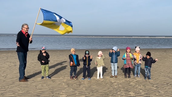 Acht Kleinkinder stehen mit einem Erwachsenen, der eine große Ukraineflagge mit Friedentaube über sie wehen lässt, am Strand, im Hintergrund ist die Nordsee zu sehen. © NDR 