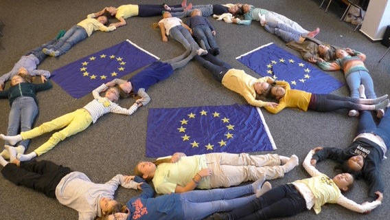 Neunzehn Mädchen liegen in Form eines Peacezeichens auf einem grauen Teppichboden, zwischen ihnen sind drei Europaflaggen ausgebreitet. © NDR 