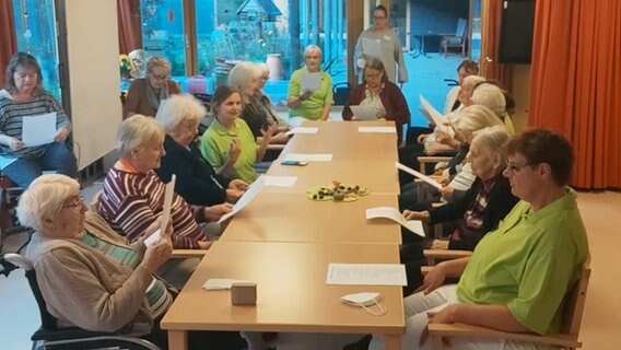 Vierzehn Frauen in Alltagskleidung, vornehmlich Seniorinnen, und drei Frauen in hellgrünen Poloshirts sitzen im Raum mit großen Fensterfronten um eine Tischgruppe, halten einen Zettel in der Hand und singen. © NDR 