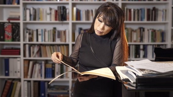 Die Komponistin Unsuk Chin steht vor einem Bücherregal und Blättert in einem Notenbuch. © dpa Bildfunk Foto: Rui Camilo