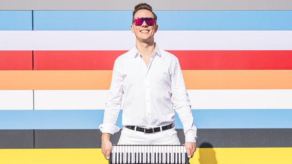 Ein lächelnder Mann mit Sonnenbrille und weißem Hemd hält ein Akkordeon mit beiden Händen vor sich. Der Hintergrund ist bunt gestreift. Es ist der Akkordeonist Martynas Levickis. © Sebastian Madej 