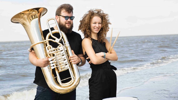 Ein Mann mit Tuba und eine Frau mit Schlagzeugstöcken stehen an einem Strand. © Norbert Schnorrenberg 