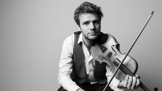 Ein junger Mann sitzt vor einer Wand und hält seine Geige am Kinn, bereit zum Spielen. © Matthias Well 