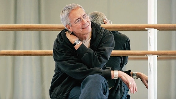 John Neumeier sitzt im Ballettsaal vor Ballettspiegel mit Ballettstangen © Collection Rolf Heyne 