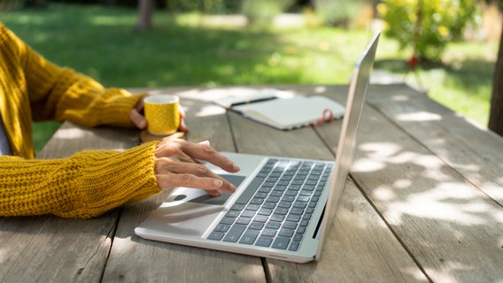 Der Arm einer Frau bedient einen Laptop, der auf einem Tisch in einem Garten steht, während die andere Hand einen Becher hält. © picture alliance / Westend61 | Svetlana Karner 