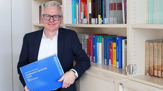 Ein Mann hält ein dickes blaues Buch in der Hand und lächelt in die Kamera. © Christian-Albrechts-Universität zu Kiel Foto: Frederike Köhl