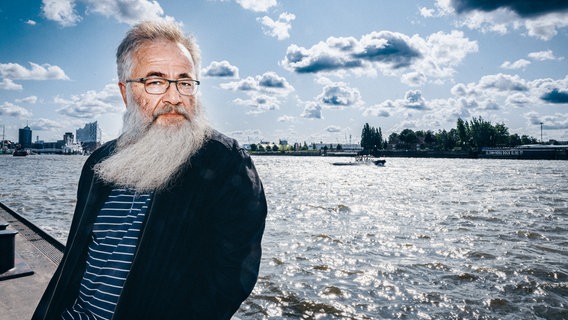 Ein älterer Herr mit grauem Wuschelbart blickt nachdenklich durch seine Brille in die Kamera. Im Hintergrund sind Wasser und die Elbphilharmonie zu sehen. Die Sonne scheint, der Himmel ist Blau und hängt volle Wolken. Der Wind kommt von rechts und lässt den Bart nach links wehen. © NDR Foto: Andreas Rehmann