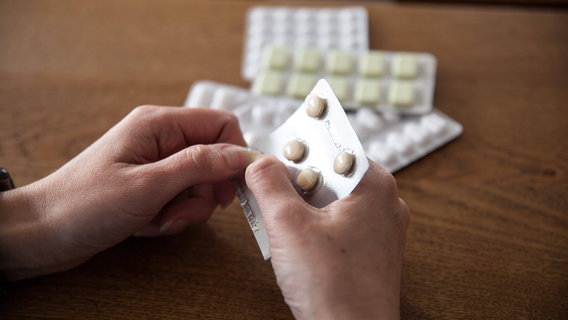 Zwei Hände halten einen Tabletten-Blister und drücken eine Pille heraus. Im Hintergrund liegt ein Stapel weiterer unterschiedlicher Paletten. © IMAGO / Fotostand 