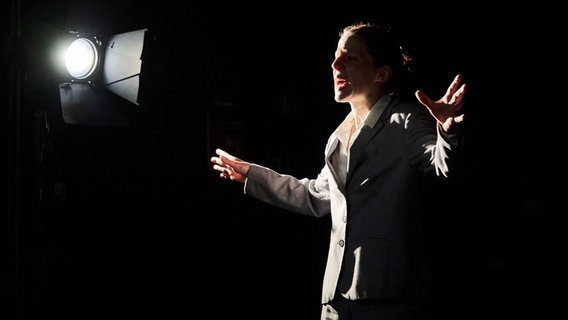 Eine junge Frau im grauen Anzug steht auf der Bühne im Licht eines einzelnen Scheinwerfers und schreit. © Landestheater Schleswig-Holstein Foto: Hernik Matzen