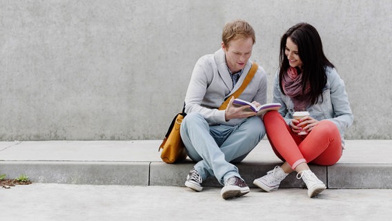 Junges Paar liest ein Buch sitzend auf einer Bordsteinkante © imago/Westend61 