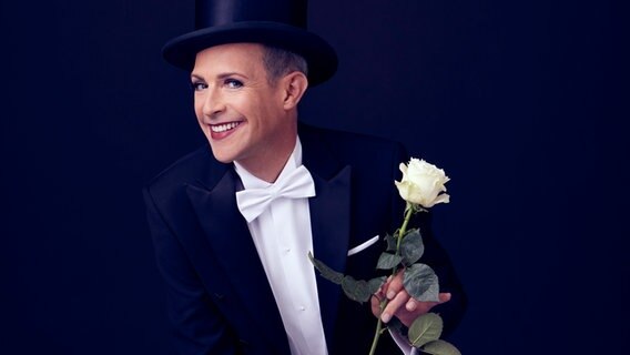 Tim Fischer, in Konzertgarderobe, lächelt mit einer weißen Rose in der Hand in die Kamera. © Benno Kraehahn Foto: Benno Kraehahn