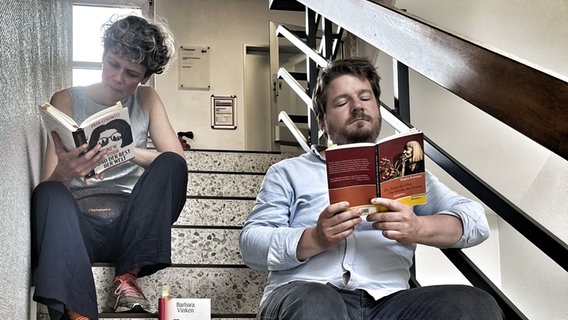 Lisa Kreißler und Alexander Solloch sitzen lesend auf Stufen eines Treppenhauses. © NDR Foto: Julia Beyer
