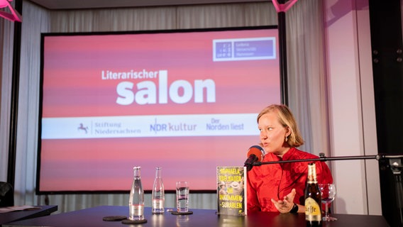 Raphaela Edelbauer sitzt an einem Tisch auf einer Bühne und spricht. © Lasse Branding Foto: Lasse Branding