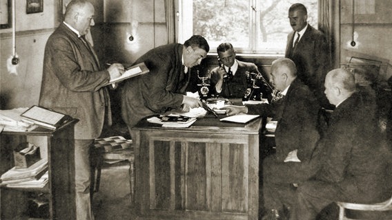 Auf einem Schwarz-Weiß-Bild steht und sitzt eine Gruppe Männer an um einen Schreibtisch herum. © obs/ZDFinfo ZDF/rbb/Polizeihistorische Sammlung 
