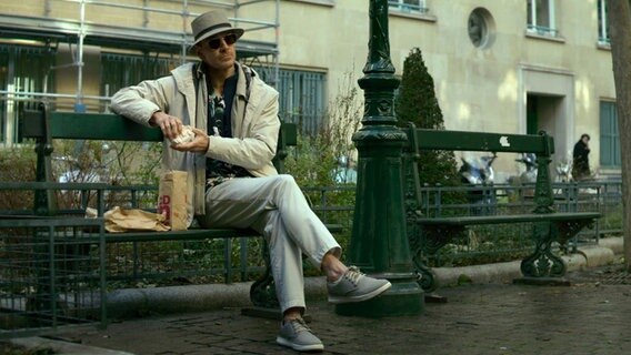 Ein Mann mit Fischerhut sitzt mit überschlagenen Beinen auf einer Bank - Szene aus "The Killer" von David Fincher © Netflix 