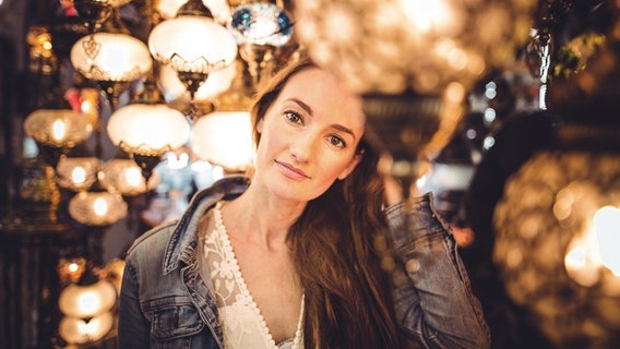 Eine junge Frau mit langem Haar steht zwischen vielen leuchtenden, orientalischen Lampen. Sie trägt eine weiße Spitzenbluse und eine Jeansjacke. Sie blickt verträumt und nachdenklich in die Kamera. © Canan Uzerli Foto: Lüey Nohut