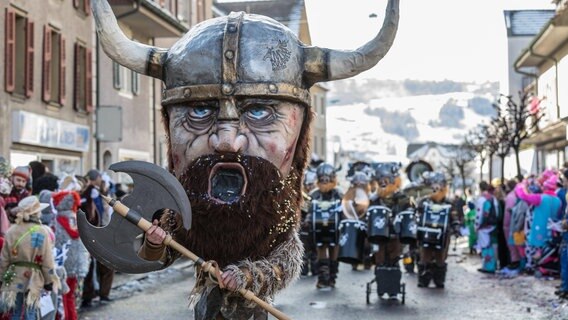 Auf einer Straßenparade läuft ein Mensch mit einer riesigen Wikinger-Maske inklusive behörntem Wikingerhelm aus Pappmaschee. er träht eine große Doppelaxt in der Hand. © IMAGO / imagebroker 