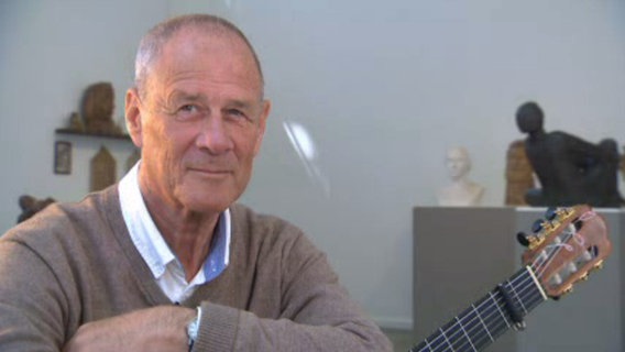 Der Liedermacher Wolfgang Rieck sitzt mit seiner Gitarre in einem Raum mit Skulpturen. © NDR 