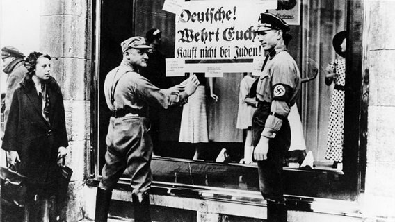 SA-Männer kleben ein Plakat mit der Aufschrift 'Deutsche! Wehrt Euch! Kauft nicht bei Juden' an die Schaufensterscheibe eines jüdischen Geschäfts. © picture-alliance / dpa Foto: dpa