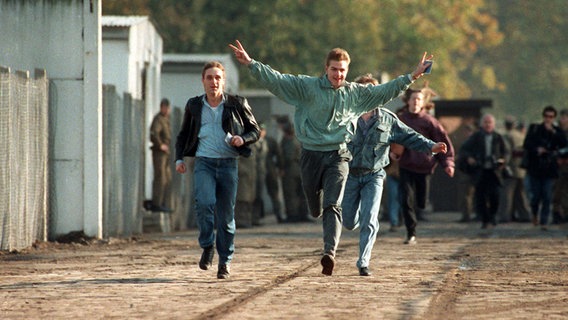 Jubelnd laufen drei junge Ost-Berliner am 10. November 1989 durch einen Berliner Grenzübergang. © dpa 