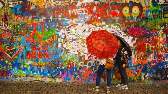 Zwei Menschen mit Regenschirmen vor einer bunt mit Schriftzeichen und Symbolen bemalten Wand © picture alliance/chromorange 