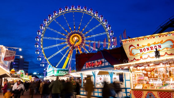 Besucher gehen über den Winterdom auf dem Hamburger Heiligengeistfeld. Im Hintergrund ist das beleuchtete Riesenrad zu sehen. © picture alliance / dpa Foto: Daniel Bockwoldt