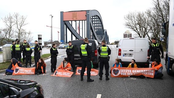 Klimaaktivisten der Bewegung "Letzte Generation" blockieren in Hamburg die Elbbrücken stadteinwärts. Vor ihnen stehen Polizisten, hinter ihnen zwei Transporter. © picture alliance / dpa Foto: Jonas Walzberg