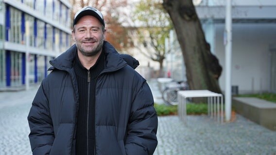 Sasha steht auf dem NDR Gelände am Rothenbaum und lächelt in die Kamera.  Foto: NDR/Frederike Burgdorf