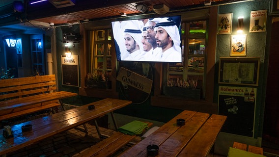 Leere Sitzbänke und Tische im Außenbereich einer Kneipe, während auf einem Fernseher das Eröffnungsspiel der WM in Katar übertragen wird. © picture alliance/dpa | Boris Roessler Foto: Boris Roessler