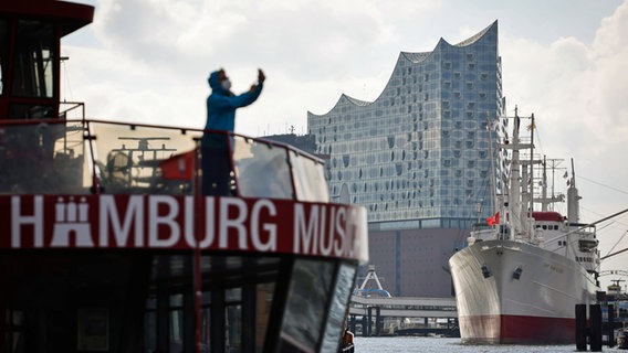 Vor der Silhouette der Elbphilharmonie und dem Museumsschiff "Cap San Diego" macht eine Frau an Deck einer Hafenfähre ein Foto mit ihrem Smartphone. © picture alliance / dpa Foto: Christian Charisius