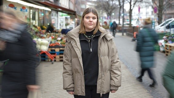 Eva guckt starr in die Kamera, während um sie herum Menschen ihren Geschäften nachgehen. © NDR Foto: Lisanne Drägert