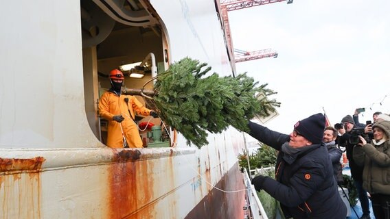 Eine Frau wirft einen Weihnachtsbaum zu zwei Seeleuten in die Luke eines Containerschiffes im Hamburger Hafen. © picture alliance / dpa Foto: Christian Charisius