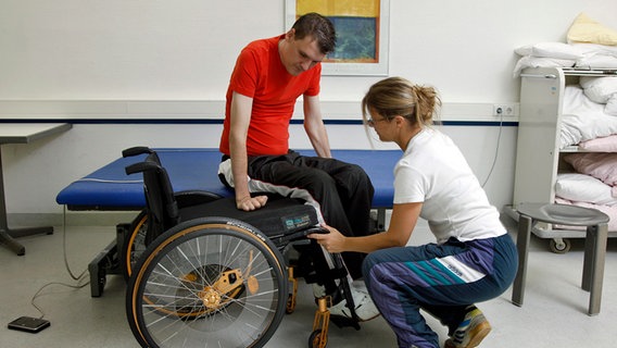 Ein bewegungseingeschränkter Mann auf einer Behandlungsliege, davor ein Rollstuhl und eine Praxismitarbeiterin. © picture alliance Foto: Jochen Tack