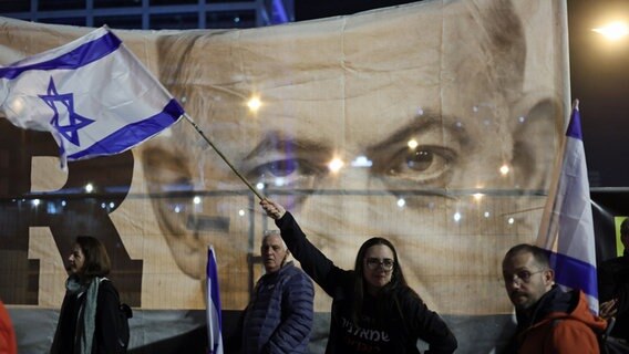 Demonstranten in Tel Aviv schwenken israelische Flaggen während einer Kundgebung gegen die neue Regierung in Tel Aviv. Im Hintergrund ist ein Banner mit dem Antlitz von Ministerpräsident Netanjahu zu sehen. © Ilia Yefimovich/dpa 