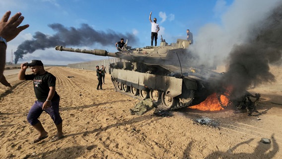Palästinenser feiern an einem zerstörten israelischen Panzer am Zaun des Gazastreifens. © Hassan Eslaiah/AP/dpa 
