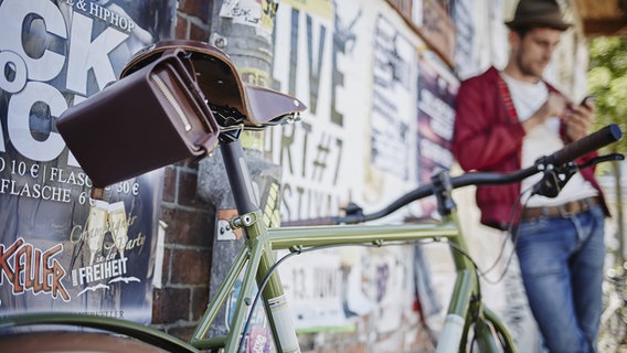 Ein junger Mann steht im Hintergrund in der Unschärfe an eine stark plakatierte Wand gelehnt, im Vordergrund steht ein sehr modernes Fahrrad. © Imago Images | Westend61 Foto: Westend61