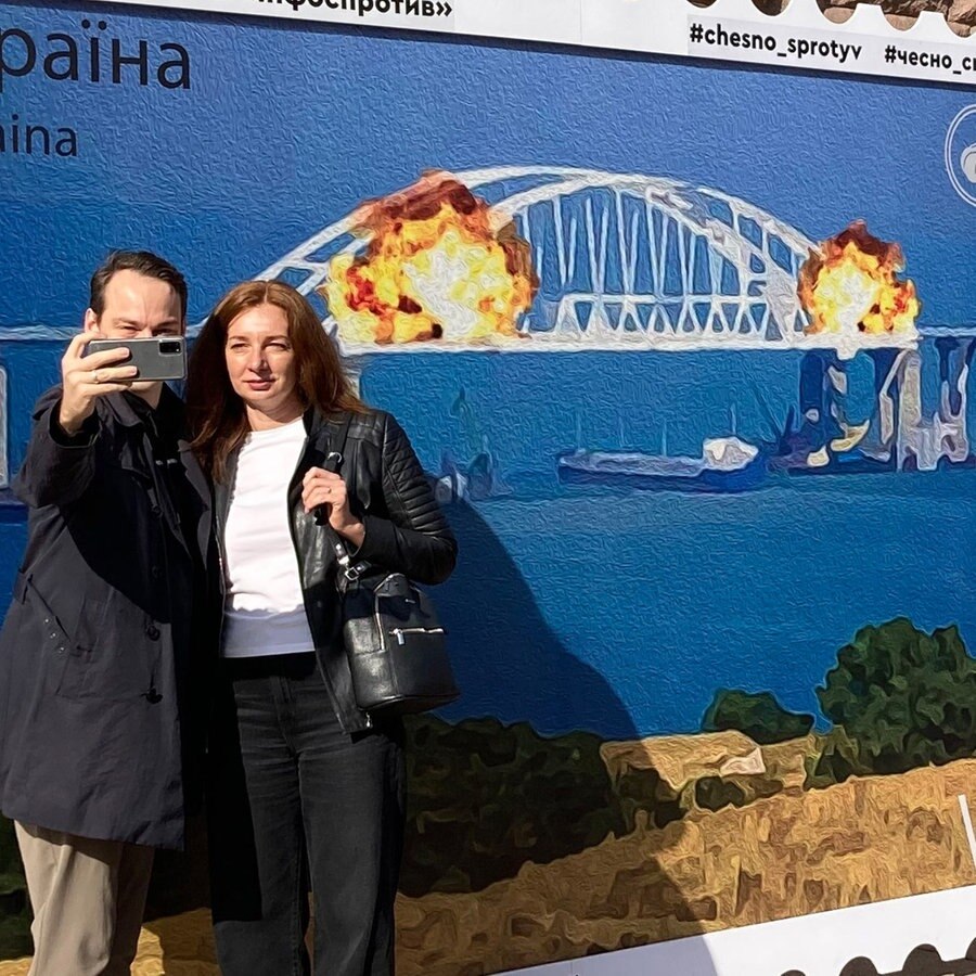 Menschen fotografieren sich in Kiew vor einem Plakat, das Explosionen auf der für Russland wichtigen Krim-Brücke zeigt. © picture alliance Foto: Hannah Wagner